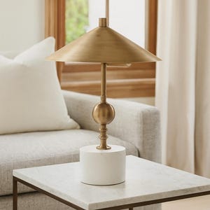 K&ouml;nnte beinhalten: Messing-Tischlampe mit konischem Schirm und wei&szlig;em Marmorsockel. Die Lampe hat einen kugelf&ouml;rmigen Akzent und einen schlanken Stiel. Die Lampe steht auf einem Tisch mit Marmorplatte.