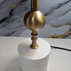 K&ouml;nnte beinhalten: Eine moderne Tischlampe mit einem wei&szlig;en, zylindrischen Sockel und einem messingfarbenen Metallstiel mit kugelf&ouml;rmigem Akzent. Das Design der Lampe ist minimalistisch und f&uuml;r verschiedene Einrichtungsstile geeignet. Die Lampe wird auf einer wei&szlig;en Oberfl&auml;che gezeigt.
