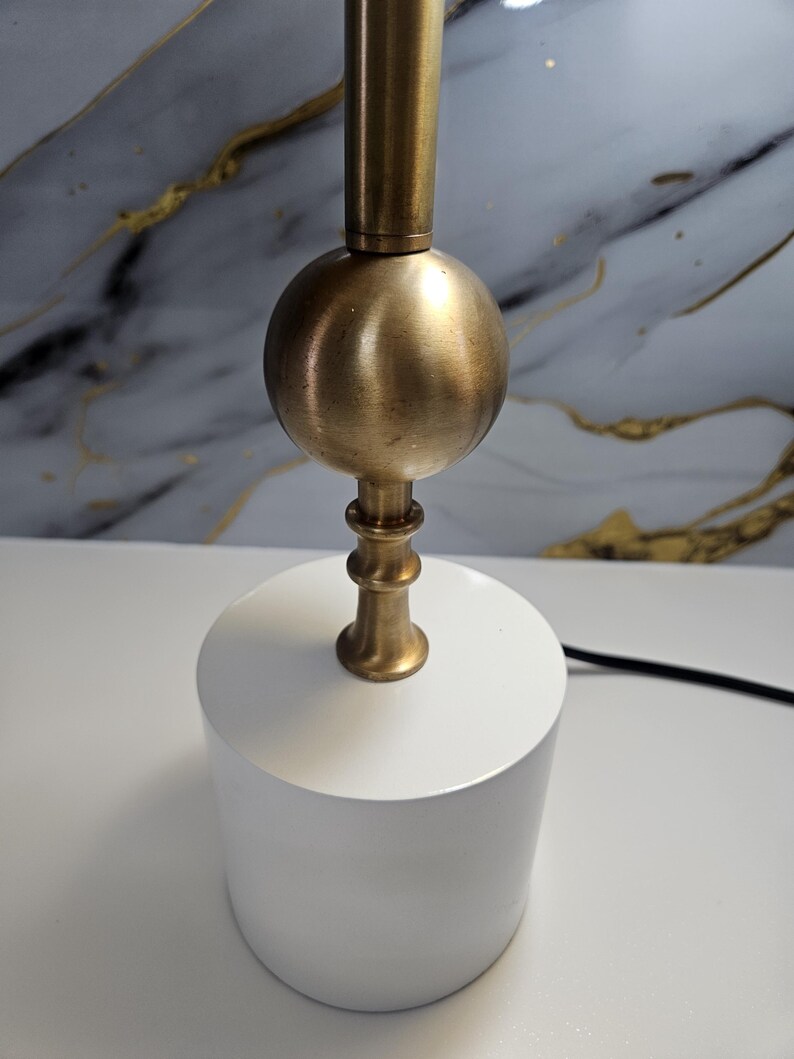 K&ouml;nnte beinhalten: Eine moderne Tischlampe mit einem wei&szlig;en, zylindrischen Sockel und einem messingfarbenen Metallstiel mit kugelf&ouml;rmigem Akzent. Das Design der Lampe ist minimalistisch und f&uuml;r verschiedene Einrichtungsstile geeignet. Die Lampe wird auf einer wei&szlig;en Oberfl&auml;che gezeigt.