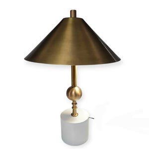 K&ouml;nnte beinhalten: Eine moderne Tischlampe mit einem konischen Messingschirm und einem Messingstiel mit kugelf&ouml;rmigem Akzent. Die Lampe steht auf einem wei&szlig;en, zylindrischen Sockel. Die Lampe ist f&uuml;r den Innenbereich konzipiert.