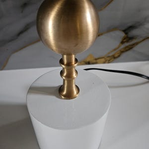 K&ouml;nnte beinhalten: Eine moderne Tischlampe mit einem zylindrischen, wei&szlig;en Sockel und einem kugelf&ouml;rmigen, messingfarbenen Oberteil. Der Lampenschaft hat ein dekoratives, mehrstufiges Design. Ein schwarzes Netzkabel erstreckt sich von der Basis.