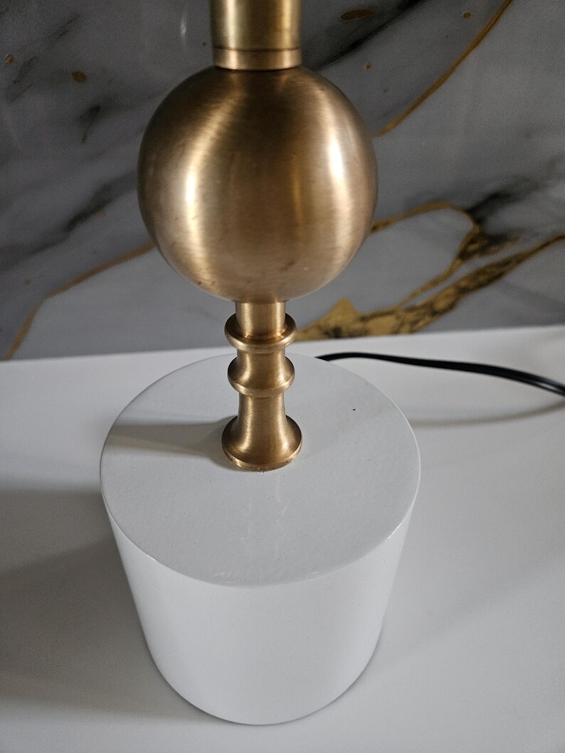 K&ouml;nnte beinhalten: Eine moderne Tischlampe mit einem zylindrischen, wei&szlig;en Sockel und einem kugelf&ouml;rmigen, messingfarbenen Oberteil. Der Lampenschaft hat ein dekoratives, mehrstufiges Design. Ein schwarzes Netzkabel erstreckt sich von der Basis.