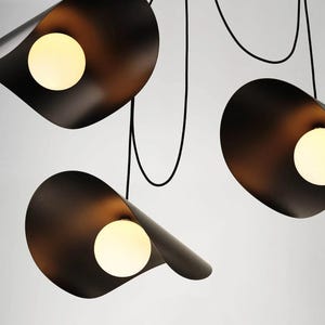 May include: Contemporary black pendant light with three petal-shaped shades. Each shade houses a warm, spherical light bulb. The fixture is suspended by black cords against a white backdrop, offering a modern design.