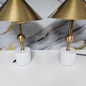 K&ouml;nnte beinhalten: Zwei passende Tischlampen. Jede Lampe hat einen goldfarbenen konischen Schirm, einen goldfarbenen Stiel mit kugelf&ouml;rmigem Akzent und einen wei&szlig;en zylindrischen Sockel. Die Lampen stehen auf einer wei&szlig;en Oberfl&auml;che mit Marmormuster im Hintergrund.