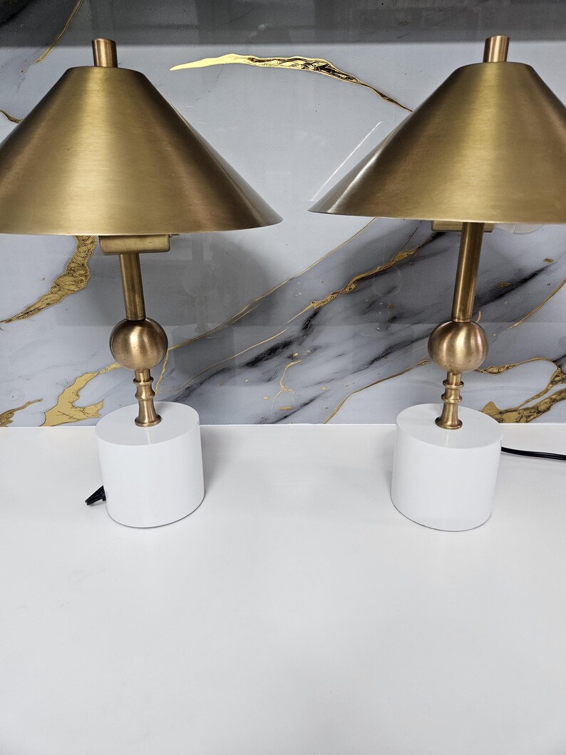 K&ouml;nnte beinhalten: Zwei passende Tischlampen. Jede Lampe hat einen goldfarbenen konischen Schirm, einen goldfarbenen Stiel mit kugelf&ouml;rmigem Akzent und einen wei&szlig;en zylindrischen Sockel. Die Lampen stehen auf einer wei&szlig;en Oberfl&auml;che mit Marmormuster im Hintergrund.