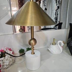 K&ouml;nnte beinhalten: Eine goldfarbene Schreibtischlampe mit konischem Schirm und wei&szlig;em zylindrischen Sockel. Die Lampe hat einen kugelf&ouml;rmigen Akzent und einen schlanken Stiel. Ein schwanf&ouml;rmiger Seifenspender ist ebenfalls zu sehen.