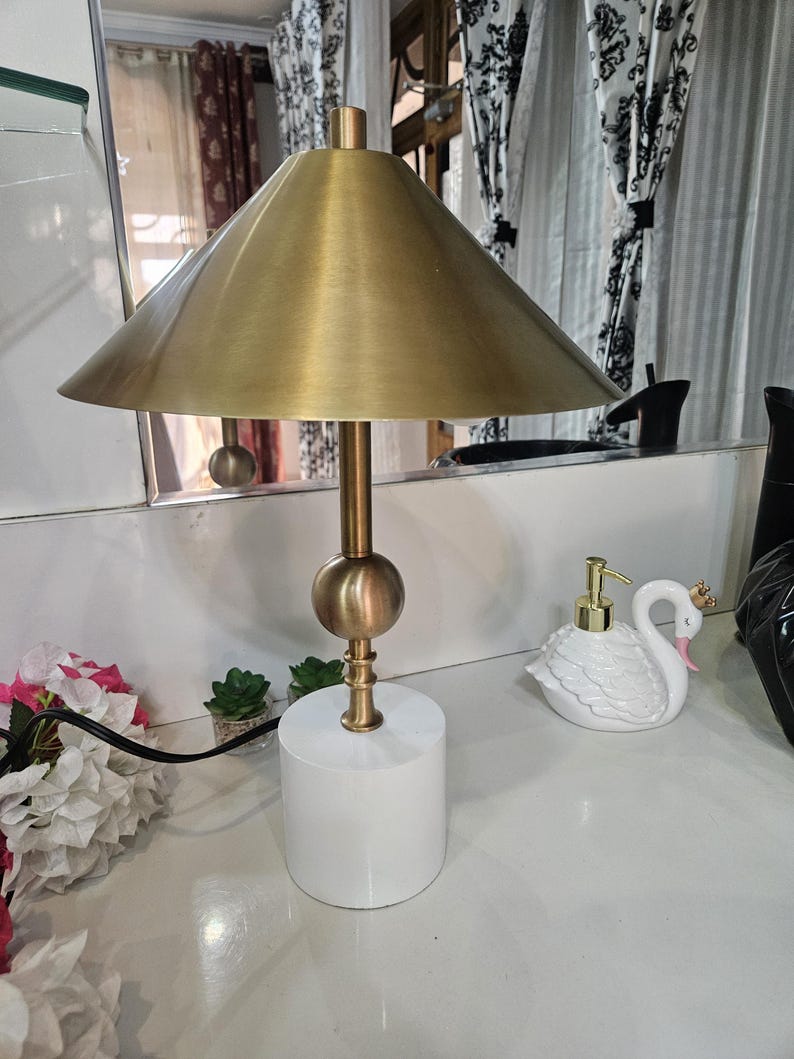 K&ouml;nnte beinhalten: Eine goldfarbene Schreibtischlampe mit konischem Schirm und wei&szlig;em zylindrischen Sockel. Die Lampe hat einen kugelf&ouml;rmigen Akzent und einen schlanken Stiel. Ein schwanf&ouml;rmiger Seifenspender ist ebenfalls zu sehen.