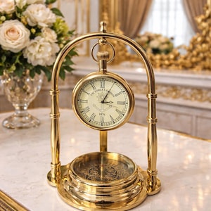 Puede incluir: Un reloj de escritorio dorado con esfera redonda, números romanos y la inscripción "Victoria London 1876". El reloj está sostenido por un soporte curvo y una base con un diseño de brújula ornamentado. Un ramo de rosas blancas está en el fondo.