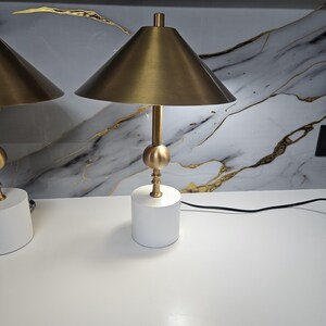 K&ouml;nnte beinhalten: Zwei passende Tischlampen mit modernem Design. Jede Lampe hat einen wei&szlig;en, zylindrischen Sockel, einen goldfarbenen Stiel mit kugelf&ouml;rmigem Akzent und einen konischen Lampenschirm. Die Lampen stehen auf einer wei&szlig;en Oberfl&auml;che vor einer marmorierten Wand.