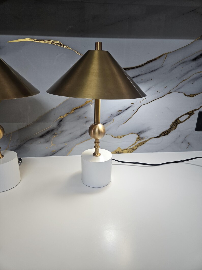 K&ouml;nnte beinhalten: Zwei passende Tischlampen mit modernem Design. Jede Lampe hat einen wei&szlig;en, zylindrischen Sockel, einen goldfarbenen Stiel mit kugelf&ouml;rmigem Akzent und einen konischen Lampenschirm. Die Lampen stehen auf einer wei&szlig;en Oberfl&auml;che vor einer marmorierten Wand.