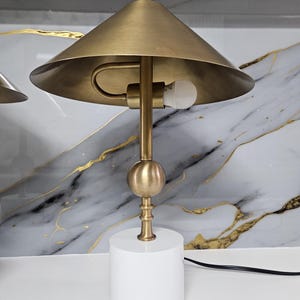 K&ouml;nnte beinhalten: Eine Tischlampe in Messingfarbe mit wei&szlig;em, zylindrischem Sockel. Die Lampe hat einen konischen Schirm und ein kugelf&ouml;rmiges Element. Der Lampenstiel ist messingfarben, und die Gl&uuml;hbirne ist sichtbar. Die Lampe ist etwa 46 cm hoch.