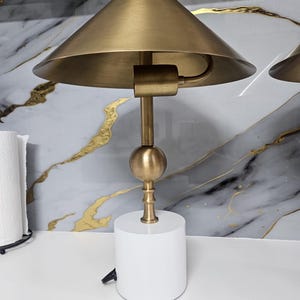 K&ouml;nnte beinhalten: Eine moderne Tischlampe mit einem wei&szlig;en, zylindrischen Sockel und einem konischen Schirm in Messingfarbe. Die Lampe hat einen Messingstiel und einen kugelf&ouml;rmigen Akzent. Das Design ist schlicht und minimalistisch.
