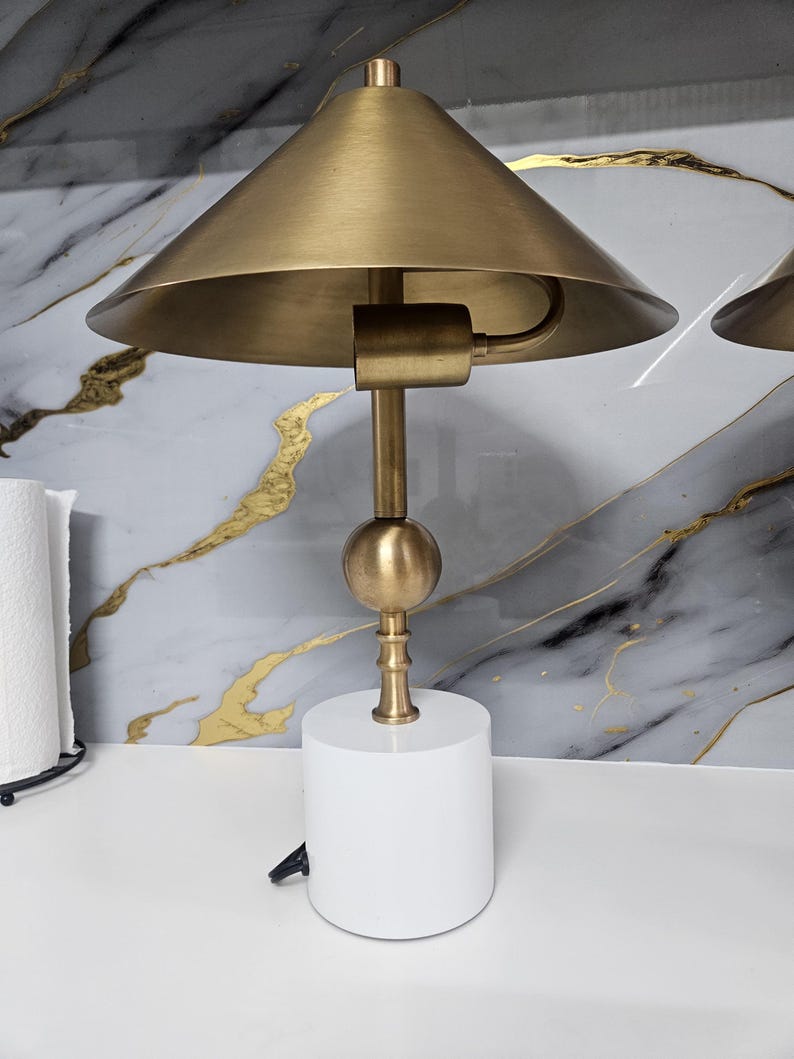 K&ouml;nnte beinhalten: Eine moderne Tischlampe mit einem wei&szlig;en, zylindrischen Sockel und einem konischen Schirm in Messingfarbe. Die Lampe hat einen Messingstiel und einen kugelf&ouml;rmigen Akzent. Das Design ist schlicht und minimalistisch.