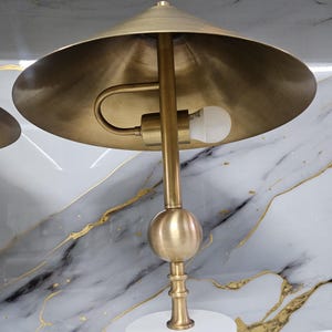K&ouml;nnte beinhalten: Messingfarbene Schreibtischlampe mit konischem Schirm und kugelf&ouml;rmigem Akzent. Die Lampe hat einen wei&szlig;en, zylindrischen Sockel und einen gebogenen Arm, der die Gl&uuml;hbirne h&auml;lt. Der Hintergrund zeigt ein wei&szlig;es und graues Marmormuster mit Goldadern.