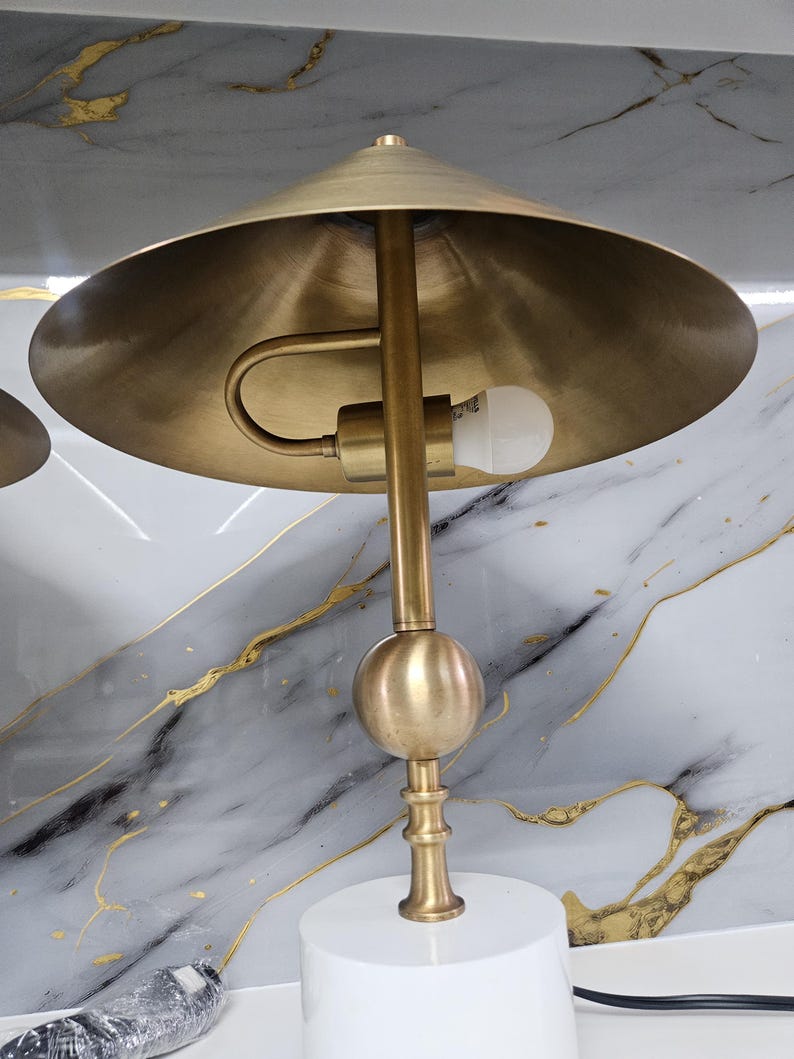 K&ouml;nnte beinhalten: Messingfarbene Schreibtischlampe mit konischem Schirm und kugelf&ouml;rmigem Akzent. Die Lampe hat einen wei&szlig;en, zylindrischen Sockel und einen gebogenen Arm, der die Gl&uuml;hbirne h&auml;lt. Der Hintergrund zeigt ein wei&szlig;es und graues Marmormuster mit Goldadern.