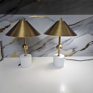 K&ouml;nnte beinhalten: Zwei passende Tischlampen. Jede Lampe hat einen goldfarbenen konischen Schirm, einen goldfarbenen Stiel mit einer dekorativen Kugel und einen wei&szlig;en zylindrischen Sockel. Die Lampen stehen auf einer wei&szlig;en Oberfl&auml;che.