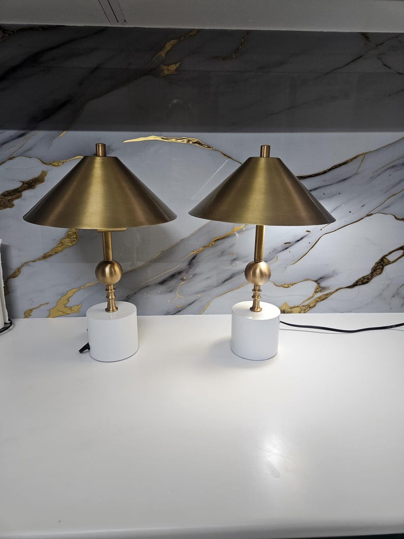 K&ouml;nnte beinhalten: Zwei passende Tischlampen. Jede Lampe hat einen goldfarbenen konischen Schirm, einen goldfarbenen Stiel mit einer dekorativen Kugel und einen wei&szlig;en zylindrischen Sockel. Die Lampen stehen auf einer wei&szlig;en Oberfl&auml;che.