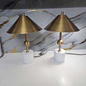 K&ouml;nnte beinhalten: Zwei passende Tischlampen mit konischen Lampenschirmen in Messingfarbe und goldfarbenen Stielen. Die Lampen stehen auf wei&szlig;en, zylindrischen Sockeln. Der Hintergrund zeigt ein marmor&auml;hnliches Muster mit goldenen Adern.