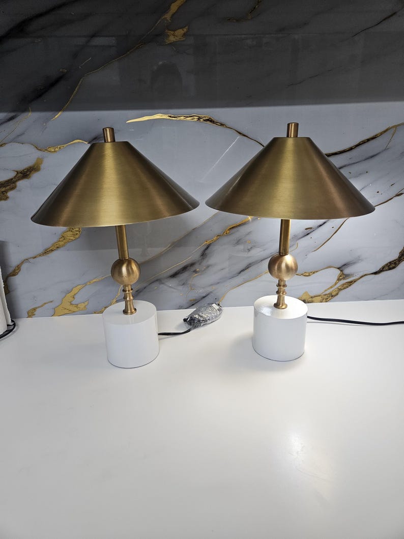 K&ouml;nnte beinhalten: Zwei passende Tischlampen mit konischen Lampenschirmen in Messingfarbe und goldfarbenen Stielen. Die Lampen stehen auf wei&szlig;en, zylindrischen Sockeln. Der Hintergrund zeigt ein marmor&auml;hnliches Muster mit goldenen Adern.