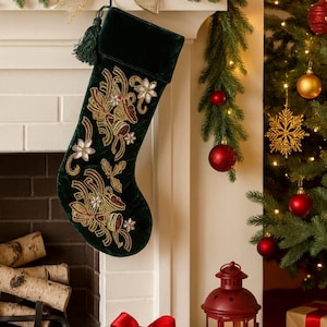 Puede incluir: Un calcetín navideño de terciopelo verde oscuro con bordados florales dorados y blancos cuelga sobre una chimenea. La escena incluye un árbol de Navidad decorado, adornos rojos y una caja de regalo con un lazo rojo.