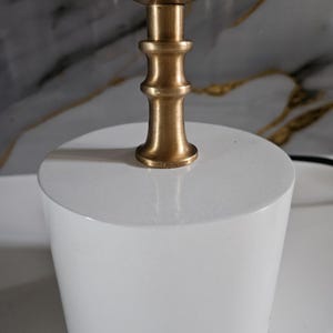 K&ouml;nnte beinhalten: Eine moderne Tischlampe mit einem wei&szlig;en, zylindrischen Sockel und einem messingfarbenen Oberteil. Der Lampenschaft hat ein dekoratives, mehrstufiges Design. Die Lampe ist f&uuml;r den Innenbereich konzipiert und bietet Umgebungslicht.