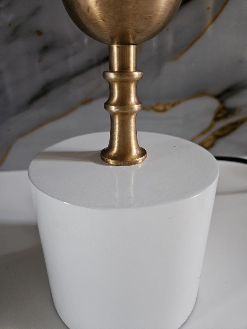 K&ouml;nnte beinhalten: Eine moderne Tischlampe mit einem wei&szlig;en, zylindrischen Sockel und einem messingfarbenen Oberteil. Der Lampenschaft hat ein dekoratives, mehrstufiges Design. Die Lampe ist f&uuml;r den Innenbereich konzipiert und bietet Umgebungslicht.