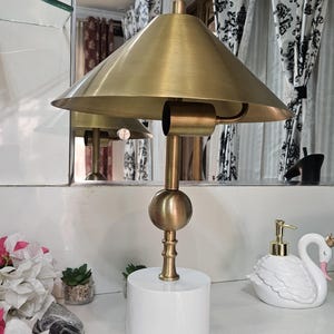 K&ouml;nnte beinhalten: Eine Schreibtischlampe in Messingoptik mit konischem Schirm und wei&szlig;em zylindrischen Sockel. Die Lampe hat einen kugelf&ouml;rmigen Akzent und einen schlanken Stiel. Die Lampe steht auf einer wei&szlig;en Oberfl&auml;che, mit einem schwanf&ouml;rmigen Seifenspender und dekorativen Blumen in der N&auml;he.