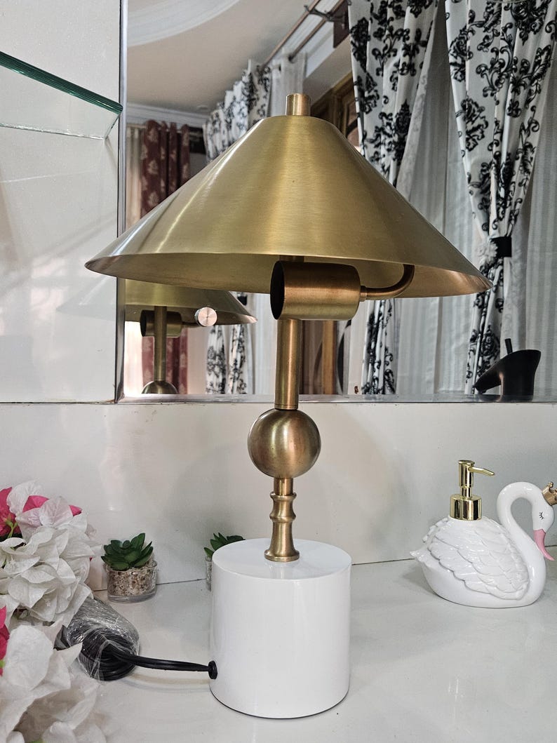 K&ouml;nnte beinhalten: Eine Schreibtischlampe in Messingoptik mit konischem Schirm und wei&szlig;em zylindrischen Sockel. Die Lampe hat einen kugelf&ouml;rmigen Akzent und einen schlanken Stiel. Die Lampe steht auf einer wei&szlig;en Oberfl&auml;che, mit einem schwanf&ouml;rmigen Seifenspender und dekorativen Blumen in der N&auml;he.
