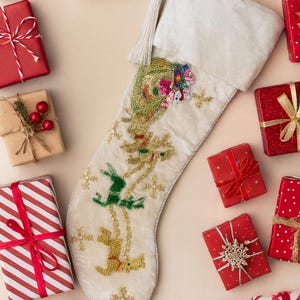 Puede incluir: Calcetín navideño crema con renos y detalles florales con cuentas. Rodeado de regalos envueltos en rojo, blanco y verde con cintas y lazos. Decoración festiva navideña.