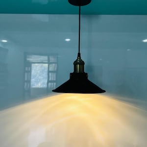 May include: Black pendant light with a conical shade and brass-coloured accents. The light hangs from the ceiling by a black cord. The light is on, emitting a warm glow.