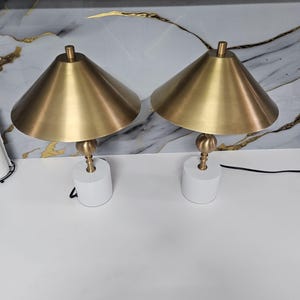 K&ouml;nnte beinhalten: Zwei passende Tischlampen. Jede Lampe hat einen wei&szlig;en, zylindrischen Sockel, einen messingfarbenen Stiel mit kugelf&ouml;rmigem Akzent und einen konischen, messingfarbenen Schirm. Die Lampen stehen auf einer wei&szlig;en Oberfl&auml;che vor einem marmorierten Hintergrund.