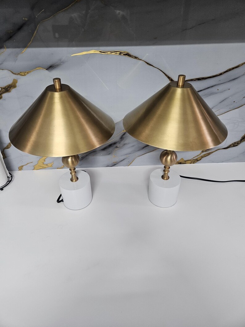 K&ouml;nnte beinhalten: Zwei passende Tischlampen. Jede Lampe hat einen wei&szlig;en, zylindrischen Sockel, einen messingfarbenen Stiel mit kugelf&ouml;rmigem Akzent und einen konischen, messingfarbenen Schirm. Die Lampen stehen auf einer wei&szlig;en Oberfl&auml;che vor einem marmorierten Hintergrund.
