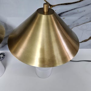 K&ouml;nnte beinhalten: Eine moderne Tischlampe mit goldfarbenem, konischem Schirm und wei&szlig;em Sockel. Die Lampe hat ein schlichtes, elegantes Design und eine Messingfarbene Spitze. Die Lampe ist mit einem schwarzen Kabel angeschlossen.