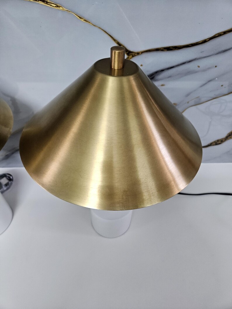 K&ouml;nnte beinhalten: Eine moderne Tischlampe mit goldfarbenem, konischem Schirm und wei&szlig;em Sockel. Die Lampe hat ein schlichtes, elegantes Design und eine Messingfarbene Spitze. Die Lampe ist mit einem schwarzen Kabel angeschlossen.