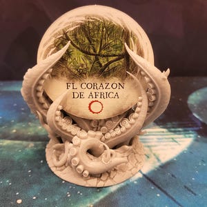 Peut inclure: Un support d'octopus en tentacules imprimé en 3D de couleur grise avec un disque rond blanc au centre. Le disque présente une image verte et brune d'une forêt avec le texte "FL CORAZON DE AFRICA" imprimé dessus.