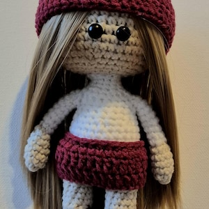 Op de afbeelding: Handgemaakte gehaakte pop met lang blond haar, met een bordeauxrode hoed en rok. De pop heeft een witte torso en armen, met zwarte knoopogen. De benen van de pop zijn wit met bordeauxrode accenten.