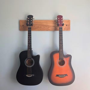 May include: Two acoustic guitars hanging on a horizontal light brown wooden wall mount. The guitar on the left is solid matte black, while the guitar on the right features a sunburst finish with a dark orange centre fading into black edges.