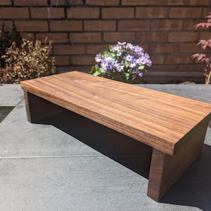 May include: A wooden riser with a natural walnut finish. The riser is made of solid wood and has a simple, modern design. It is perfect for elevating plants, books, or other decorative items.