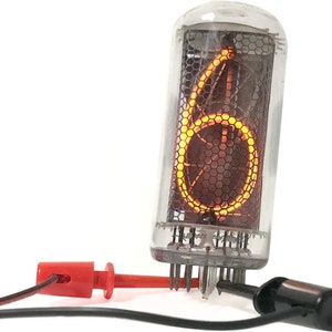 May include: A clear glass Nixie tube displaying the number "6" in a bright orange color. The tube is connected to a black and red wire with alligator clips.