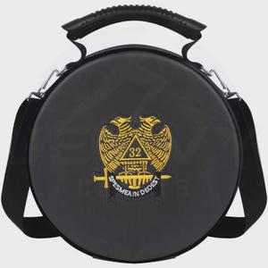 Puede incluir: Bolso redondo negro con asa superior y bandolera. El bolso presenta un emblema dorado con dos águilas, un triángulo con el número 32 y el texto "SPES MEA IN DEO EST".