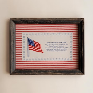 May include: A framed print featuring a red and white horizontal striped background. Centered is a white rectangular panel with a blue star border, a small American flag illustration, and the text of the Pledge of Allegiance in blue ink.