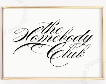 May include: A framed print with the words "The Homebody Club" in elegant black script. The text is centered on a white background, encased in a gold-colored frame. The overall aesthetic is minimalist and decorative.