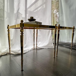 Puede incluir: Una mesa auxiliar rectangular dorada con una tapa de cristal y cuatro patas delgadas y decorativas. La mesa tiene un diseño minimalista con una pequeña vela y libros encima. La mesa está colocada frente a una ventana con cortinas blancas.