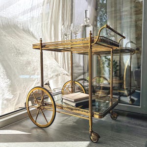 Puede incluir: Un carrito de servicio de metal dorado con dos estantes rectangulares de cristal, una barandilla decorativa y cuatro ruedas, incluyendo dos ruedas grandes de radios y dos pequeñas. El carrito sostiene un decantador de cristal y dos copas.