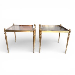 Puede incluir: Dos mesas auxiliares de latón con tapas de cristal transparente. Cada mesa tiene una tapa rectangular, patas ornamentadas y un acabado dorado. Las mesas se muestran sobre un fondo blanco.