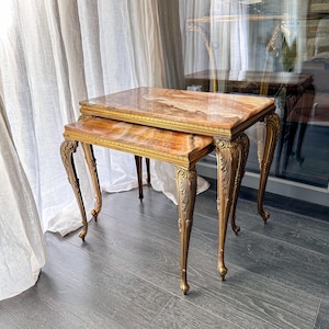 Puede incluir: Un juego de dos mesas nido con tableros rectangulares de piedra ónice pulida con vetas naranjas y marrones. Cada mesa se apoya sobre cuatro patas de metal dorado, curvas y ornamentadas, con detalles en relieve.