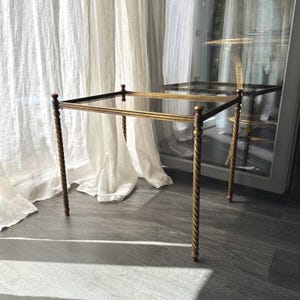 Peut inclure: Une table d'appoint rectangulaire dotée d'un plateau en verre transparent et d'un cadre en métal doré. La table repose sur quatre pieds fins et torsadés, ornés de petits embouts sphériques à chaque coin, soutenant la surface vitrée.