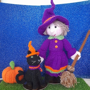Könnte beinhalten: Eine gestrickte Hexenpuppe in einem violetten Kleid und Hut mit orangefarbenen Akzenten, die einen braunen Besen hält. Neben der Puppe steht eine schwarze gestrickte Katze mit orangefarbenem Hut und lila-orangefarbenem Schal sowie ein orangefarbener Kürbis.