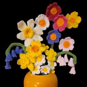 May include: A colourful display of crocheted flowers in a yellow vase. The flowers are various colours, including white, yellow, pink, red, and blue, with orange centres. The vase is a bright, glossy yellow, set against a black backdrop.