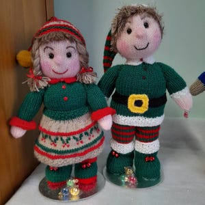 May include: Two knitted Christmas elf dolls. The dolls are wearing green and red outfits with hats. The girl elf has a green dress with a white and red skirt. The boy elf has a green top and red and green striped pants.